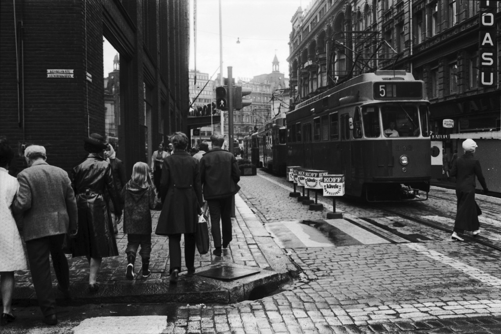 Aleksanterinkatu 52. Jalankulkijoita ylittämässä Aleksanterinkatua Keskuskadun risteyksessä Stockmannin tavaratalon kulmassa kävelykatukokeilun aikoihin. Ajoväylällä jalankulkijoille varatulla osuudella Koffin mainoksia. Oikealla kolme raitiovaunua jonossa. Näkymä Mannerheimintien suuntaan.