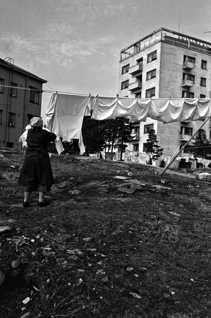 Nainen ripustaa pyykkejä narulle kuivumaan. Taustalla Mannerheimintie 118 ja Tukhomankatu 5.