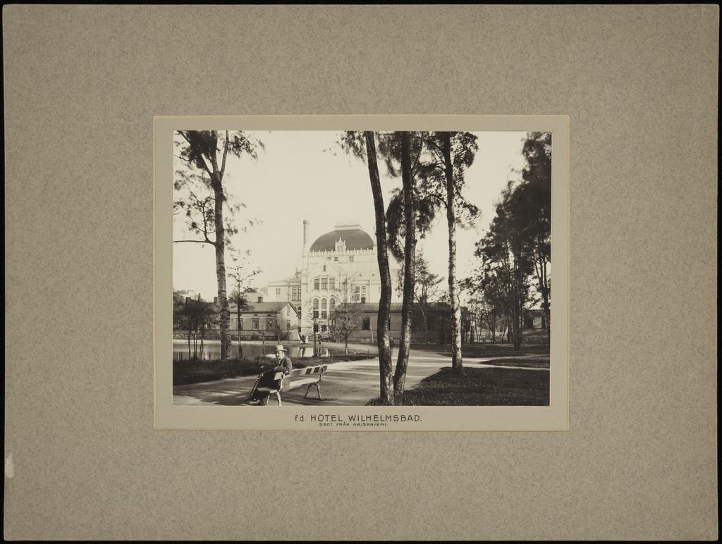 Kaisaniemen puisto, mies istuu penkillä. Vasemmalla joutsenlampi, taustalla Kansallisteatteri ja Villensaunan (Wilhelmsbad) kylpylän puurakennuksia.