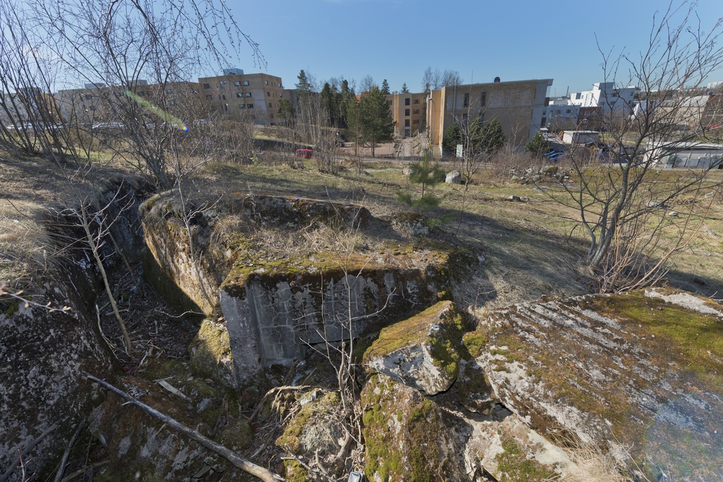 Länsimäki. Ensimmäisen maailmansodan aikainen maalinnoitus, puolustusasema II:8 Länsimäessä, Vantaalla. Kuvassa näkyy kallioon louhittua, betonilla tuettua taisteluhautaa sekä betoninen konekivääriasema aseman länsiosassa. Aseman katto on räjäytetty. Haudan puoleinen seinä on miltei kokonaan tuhoutunut. Betoninpalat täyttävät aseman ja osan haudasta. Seinässä erottuu juuri ja juuri toisen komeron yläreuna. Taustalla näkyy tie, leikkikenttä ja asuinrakennuksia. Kuvaussuuntana etelä.