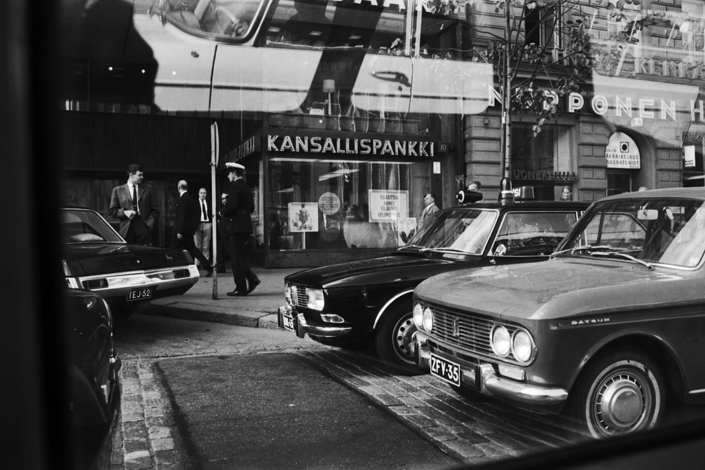 Mannerheimintie 10. Pysäköityjä autoja (vasemmalta lukien: Dodge Dart, Saab 99, Datsun Bluebird) sekä poliisi kadulla Kansallispankin (KOP) edessä Mannerheimintie 10:n kohdalla. Kuvattu bussin tai raitiovaunun ikkunasta.