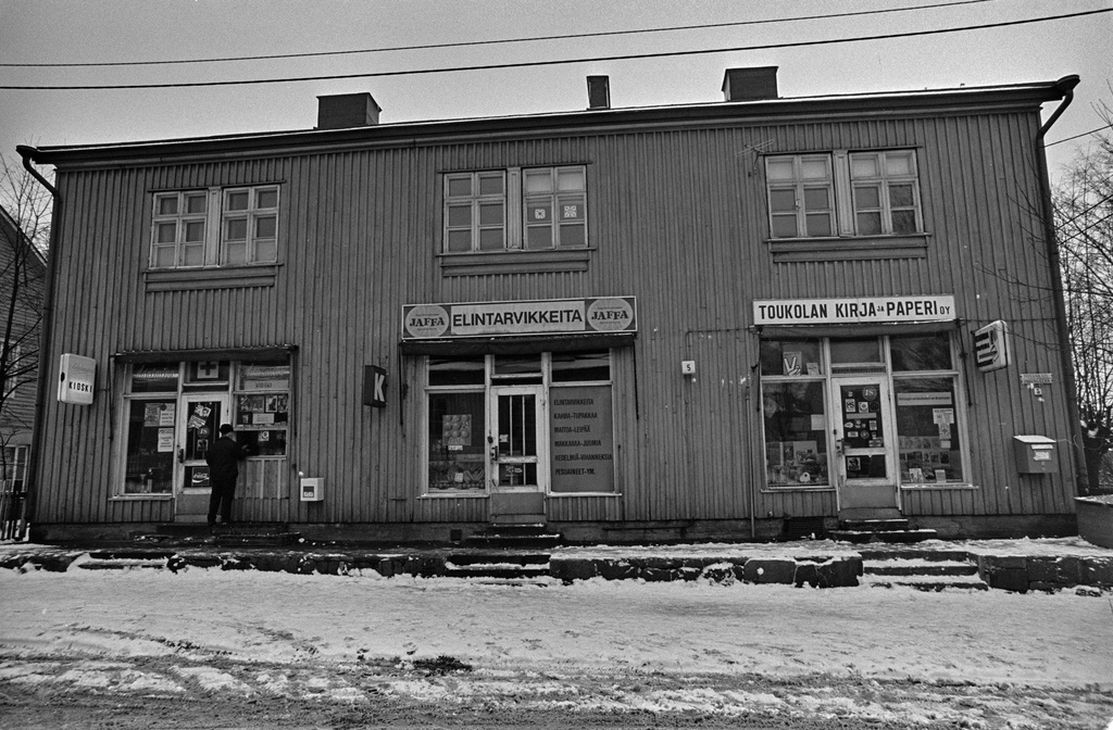 Kioski Adalmina, K-elintarvikeliike ja Toukolan kirja ja paperi Oy puurakennuksessa Toukolassa. Asiakas on asioimassa kioski Adalminan palveluluukulla.