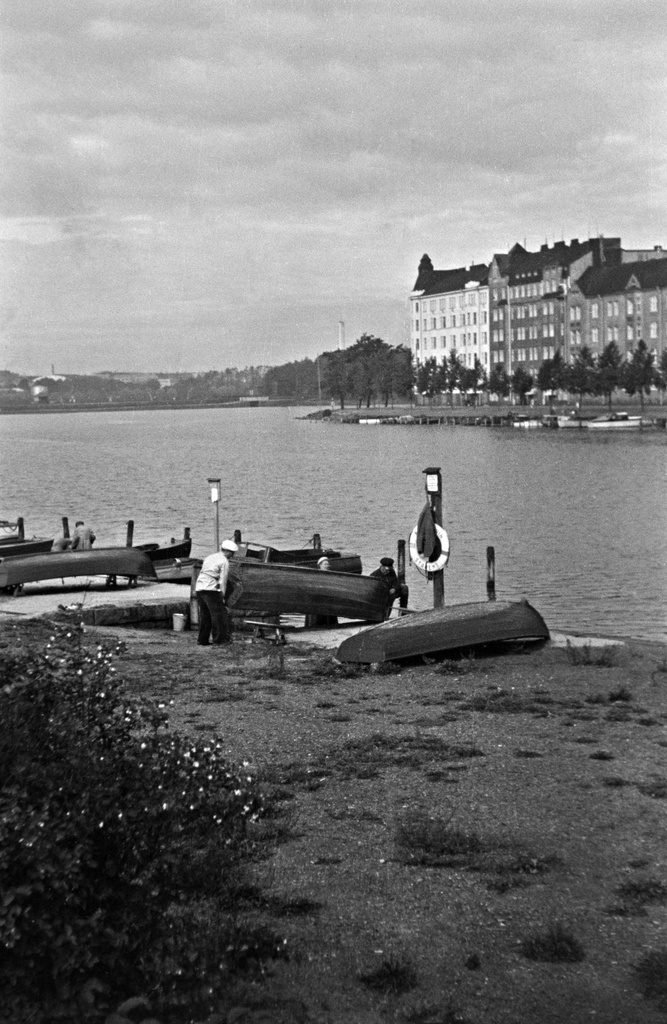 Kolme henkilöä nostamassa venettä Kaisaniemenlahden rannalla. Taustalla Pitkänsillanranta 13 - 17.