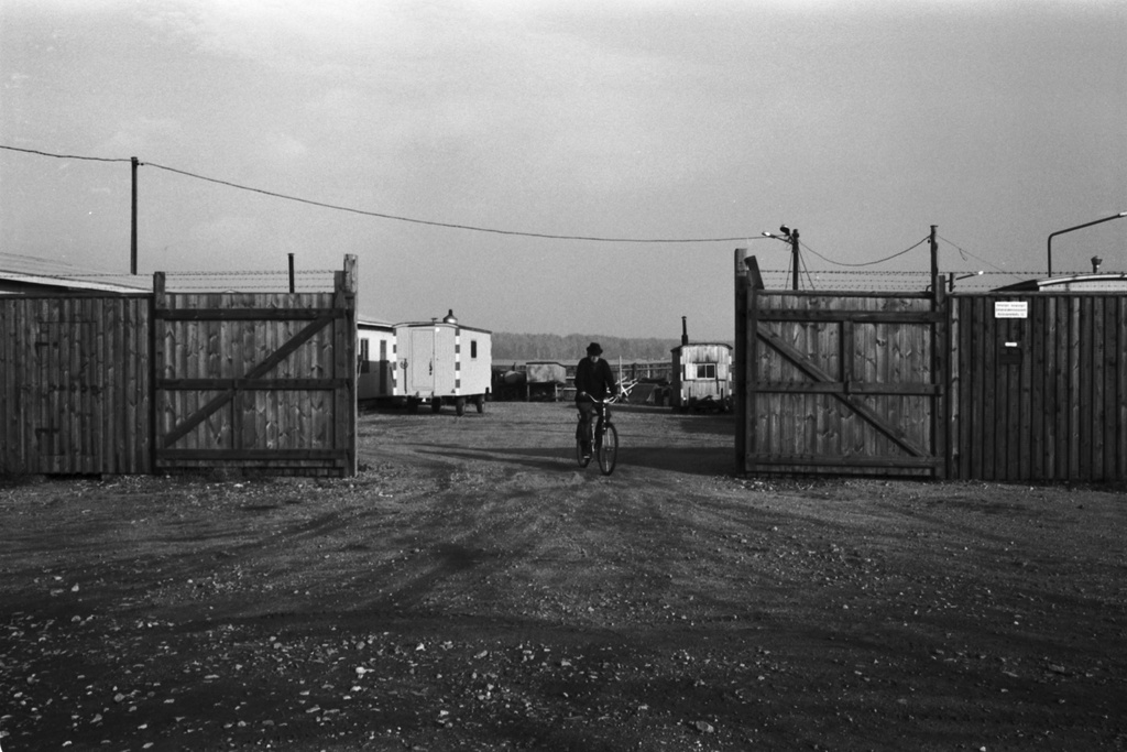 Sörnäinen. Näkymä Kyläsaarenkadun laidalta Verkkosaaresta. Mies ajaa polkupyörällä ulos Helsingin kaupungin varastoalueen lauta-aidan portista. Pihalla työmaille hinattavia tauko- ym. tupia.