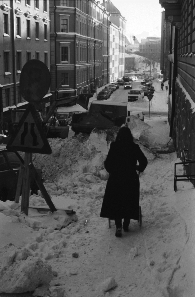 Meritullinkatu. Äiti (Etta Harvilahti) työntämässä lastenvaunuja lumikasojen keskellä Meritullinkadulla. Näkymä Rauhankadun kulmasta Kirkkokadun suuntaan.