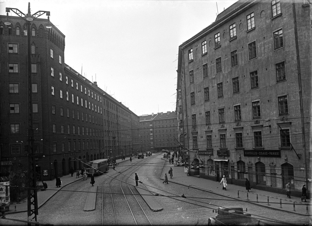 Näkymä Caloniuksenkadun ja Runeberginkadun risteyksestä Mechelininkadun suuntaan. Vasemmalla Caloniuksenkatu 9 ja 7, oikealla Caloniuksenkatu 10. Kadulla jalankulkijoita, linja-auto ja kuorma-autoja. Caloniuksenkatu 9:n rakennuksessa Elanto, Caloniuksenkatu 10:n rakennuksessa Kansallis-Osake-Pankin ja Hufvudstadsbladetin konttorit.