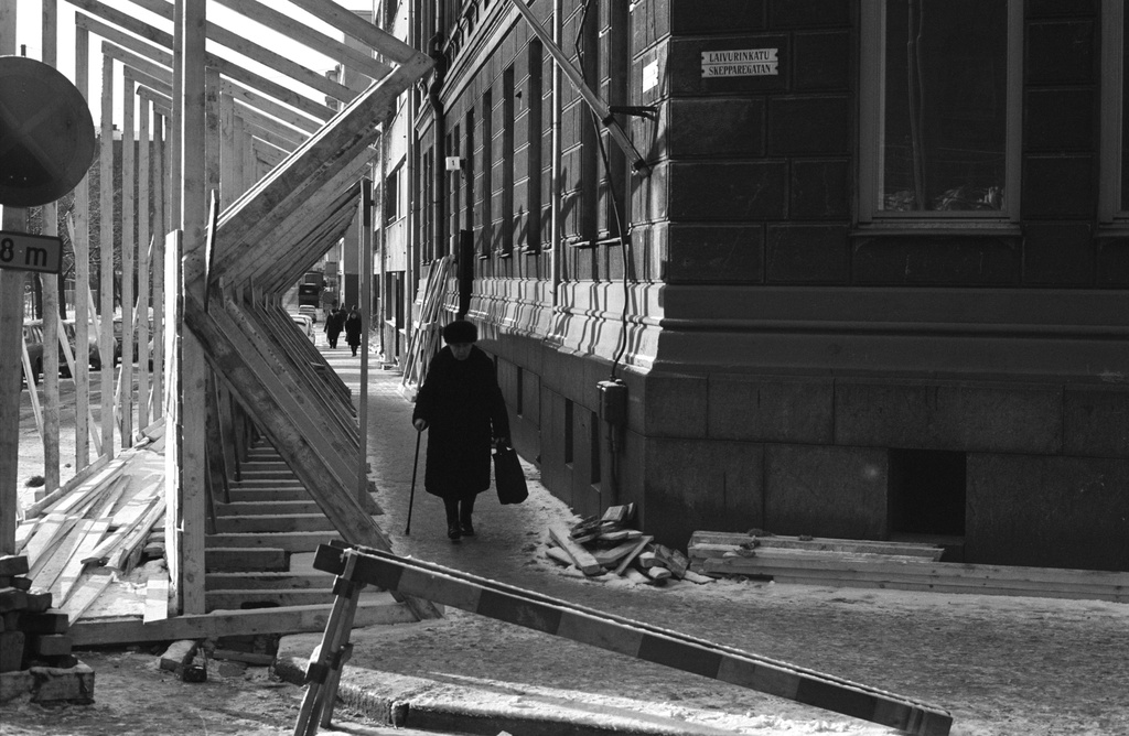 Vanha nainen kepin kanssa kävelemässä purettavan kerrostalon edustalle rakennetulla puisella jalankulkukäytävällä Laivurinkadun ja Sepänkadun kulmassa.