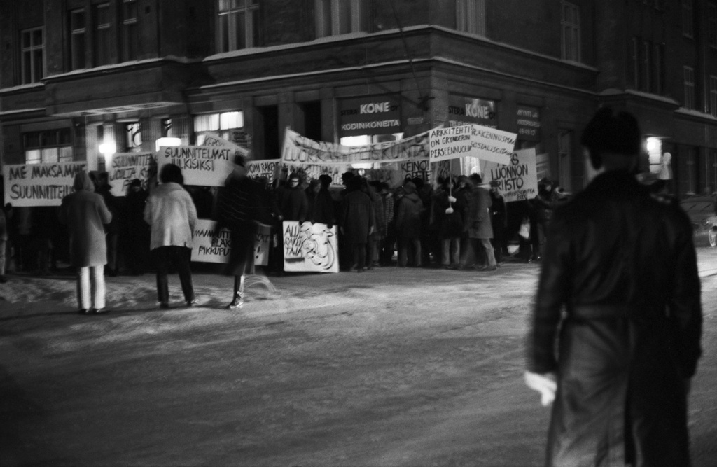 Aurorankatu 13. Nervanderinkatu 8. Mielenosoittajia plakaatteineen ja banderolleineen Aurorankadun ja Nervanderinkadun kulmassa Suomi rakentaa 4 -näyttelyn avajaisien aikaan. Etualalla, Taidehallin nurkalla seisoo poliisi. Mielenosoittajat vastustavat grynderien valtaa aluerakentamisessa.