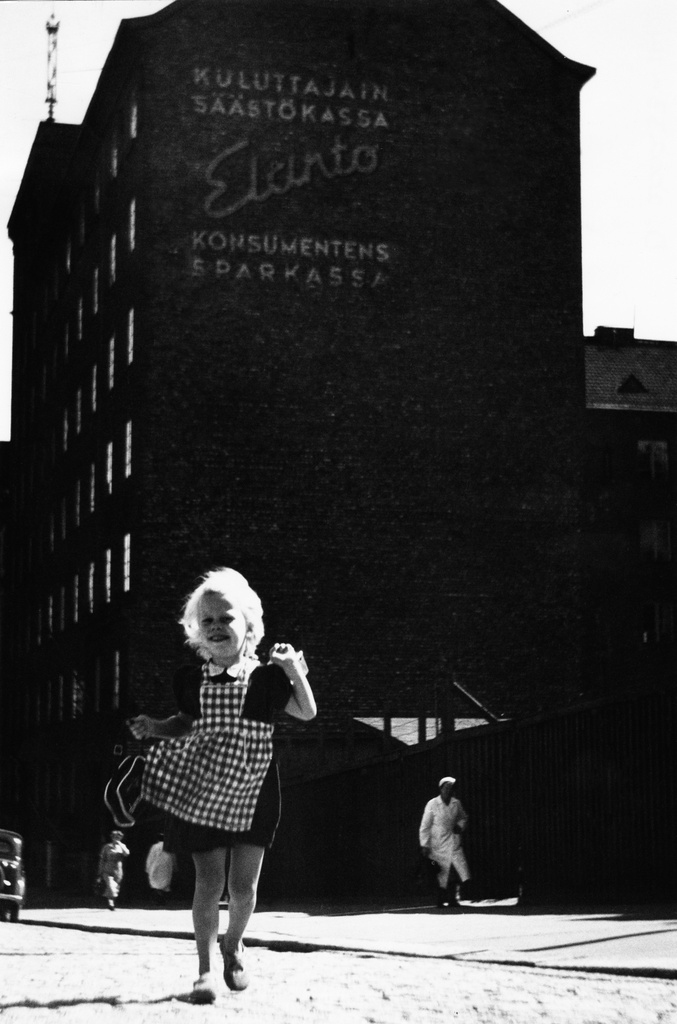 Tyttö juoksee kadulla Viidennellä linjalla Kalliossa. Taustalla Elannon asuinrakennus (Väinö Vähäkallio, 1927), sekä mainos seinässä. Elannon kokoelma.