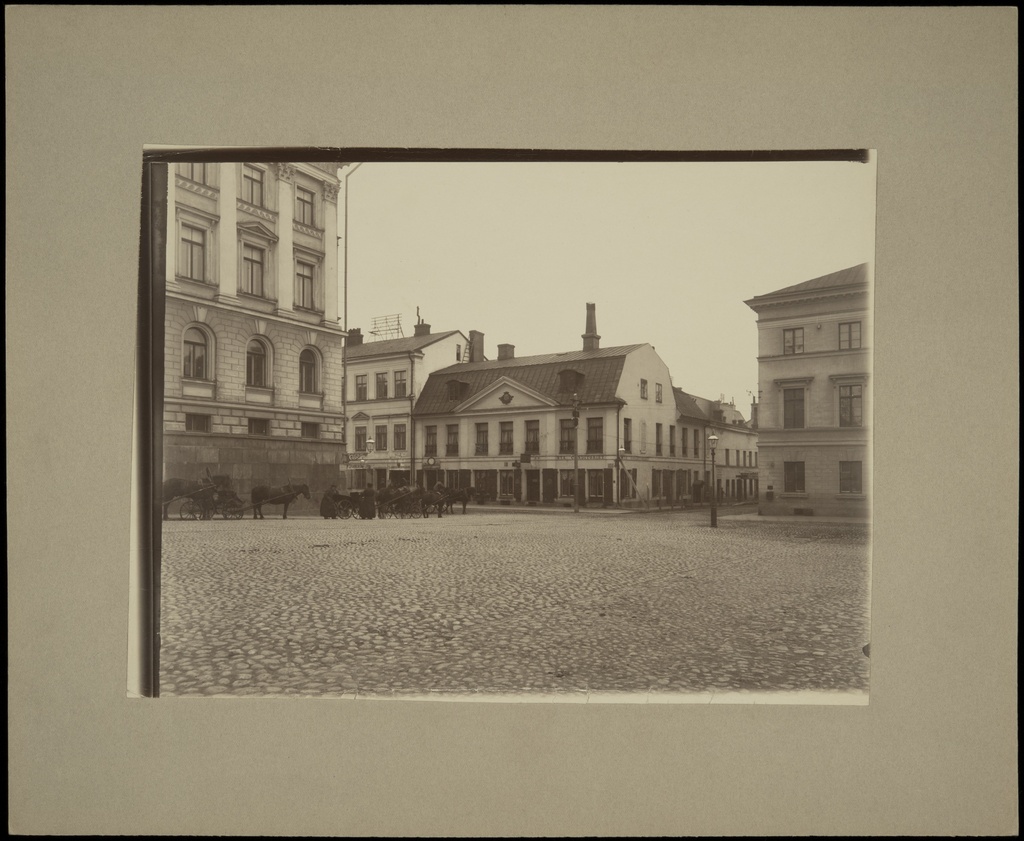 Aleksanterinkatu 18 (Katariinankatu 3) nähtynä Senaatintorilta, vasemmalla Senaatintalo (=Valtioneuvoston linna, Nikolainkatu 1, nyk. Snellmaninkatu 1) ja vuokra-ajuriasema. Aleksanterinkatu 18:ssa ajurikonttori, Lindroos & Flodströmin kelloliike sekä rouva J. Nymarkin Nya Conditoriet. Katariinankatu 3:ssa K. Löfgrenin maitokauppa.