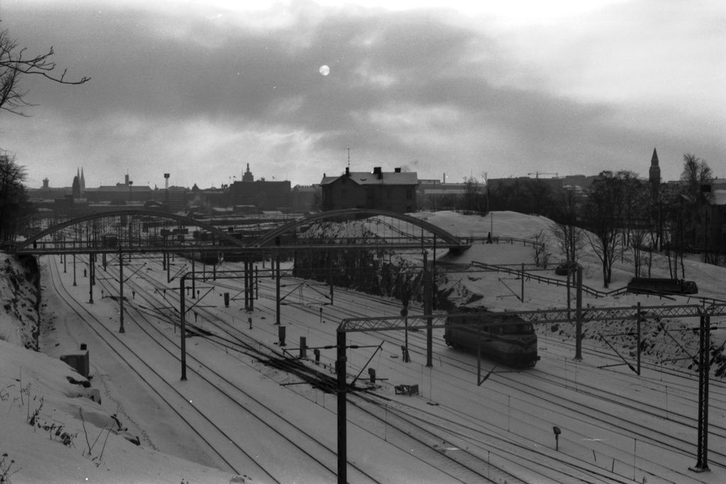 Näkymä harmaana talvipäivänä pääradan itäpuolelta etelän suuntaan.  Keskellä radan ylittävä Linnunlaulun silta, sen takana Eläintarhan huvila 12 osoitteessa Linnunlauluntie 15. Raiteilla kulussa Valtion rautateiden HR-12 -tyypin veturi. Taustalla vasemmalla mm. Helsingin rautatieaseman torni, oikealla Kansallismuseon torni.