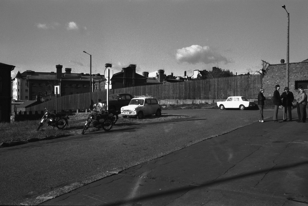 Näkymä Lutikkalinnan (Puolipäivänkatu 4) suunnalta Katajanokan vankilaan päin. Oikealla nuoria miehiä seisoskelemassa Lutikkalinnan edessä, eskellä ja vasemmalla autoja ja mopoja parkissa. Taustalla Linnankuja.