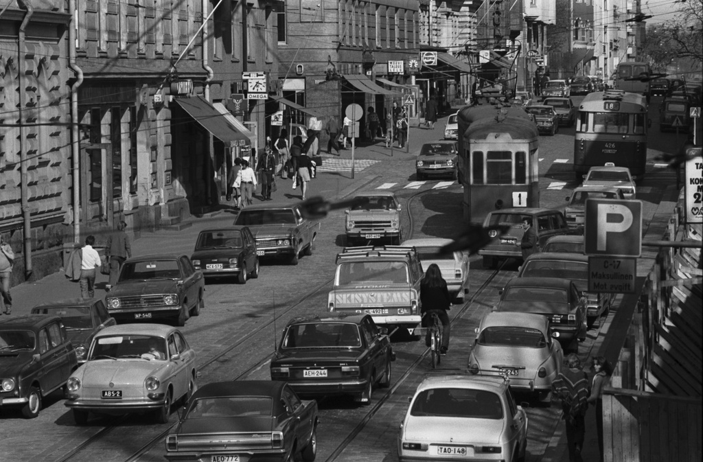 Liisankatu. Iltapäivän ruuhkaliikennettä Liisankadulla Kaisaniemen puoleisessa päässä. Etualalla polkupyöräilijä autojen keskellä. Risteyksessä linjan 1 ruuhkavuoron vanha kaksiakselinen raitiovaunu perävaunun kera ja bussi 18. Näkymä kiviaidan päältä itään Snellmaninkadun suuntaan.