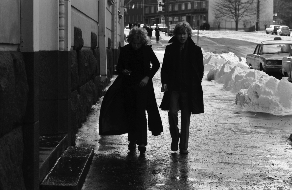 Runeberginkatu 67. Jalankulkijoita, etualalla kaksi nuorta miestä sulavan lumen kastelemalla Runeberginkadulla. Toisella maksipituinen takki. Näkymä Töölönkadun suuntaan.