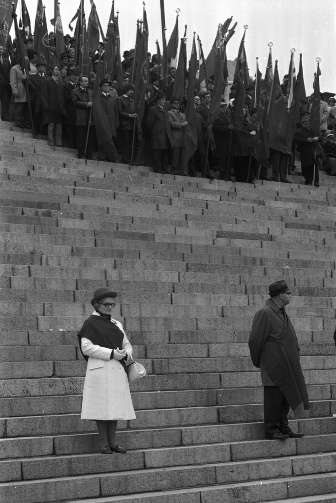 Hallituskatu 7. Kaksi ihmistä, joilla airutnauhat seisomassa Tuomiokirkon portailla työväen vappujuhlassa 1.5.1972. Ylempänä ihmisiä lippujen kanssa.