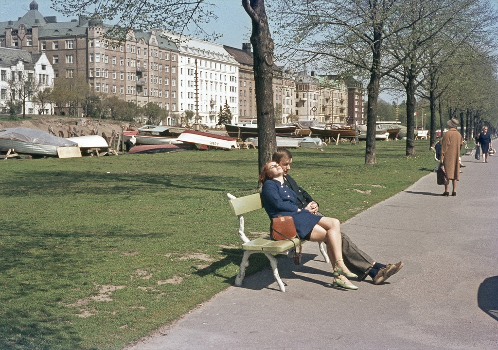 Merisatamaranta (=Merisatamanranta), nuori nainen ja mies puiston penkillä. Taustalla oikealla jalankulkijoita, vasemmalla veneitä ja Merikadun rakennuksia.