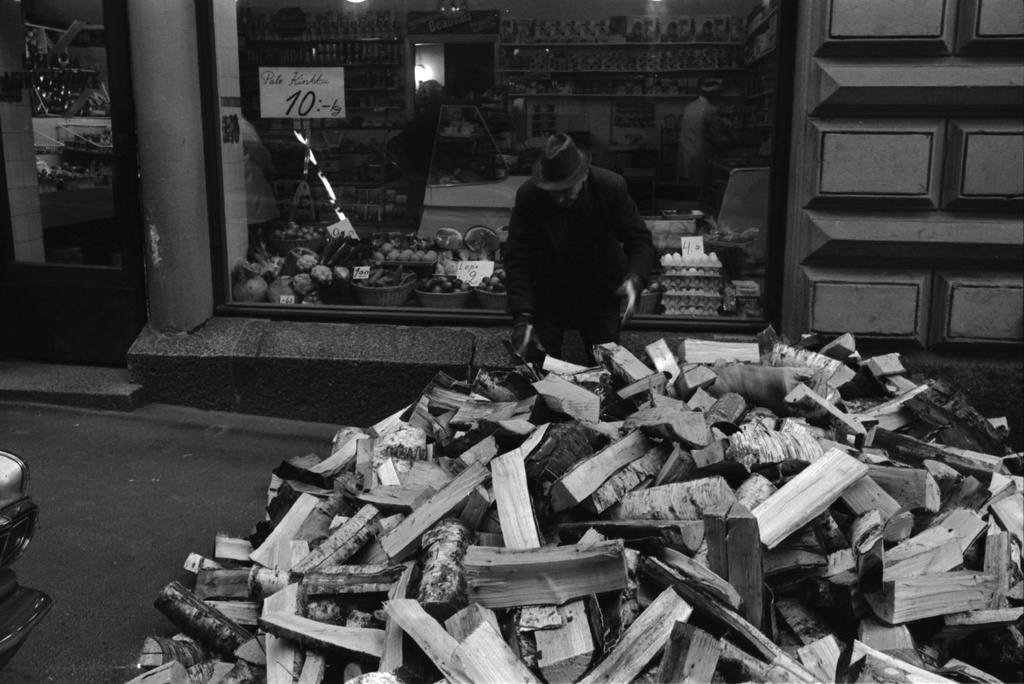 Kruununhaka, Meritullinkatu 25. Talonmies Ville Sneck latomassa jalkakäytävälle kumottuja klapeja kellarin luukusta sisään Meritullinkadulla Liisankadun kulmassa. Taustalla lihakaupan näyteikkuna. Talo Liisankatu 7 - Meritullinkatu 25 oli puulämmiteinen vuoteen 1971, jolloin talo liitettiin kaukolämpöverkkoon.