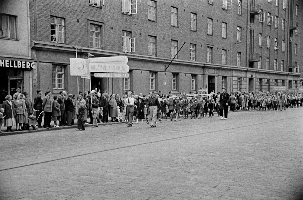 Rauhanmarssi ja yleisöä Helsinginkatu 10:n kohdalla, Suomen Demokraattisen Nuorisoliiton (SDNL) jäseniä. Lapset kantavat mm. lennokkeja sekä kylttiä "Haluamme askarrella, vaadimme rauhaa".