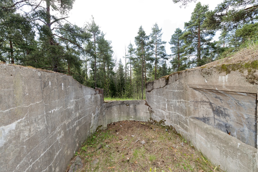 Laajalahti. Ensimmäisen maailmansodan aikainen maalinnoitus, puolustusasema XXXI:4 Laajalahdessa, Espoossa. Kuvassa on betonista rakennettu kaarevaseinäinen konekivääriasema. Aseman katto on jäänyt rakentamatta. Kuvassa oikealla seinällä on konekiväärin jäähdytysvesisäiliötä varten tehty syvennys. Ampuma-aukon vasemmalla puolella aseman sisältä katsottuna näkyy aseman tukemiseen käytettyjä teräspalkkeja. Ampuma-aukon alapuolella puolestaan erottuu syvennys konekiväärin jaloille. Aseman pohjalla kasvaa ruohoa.