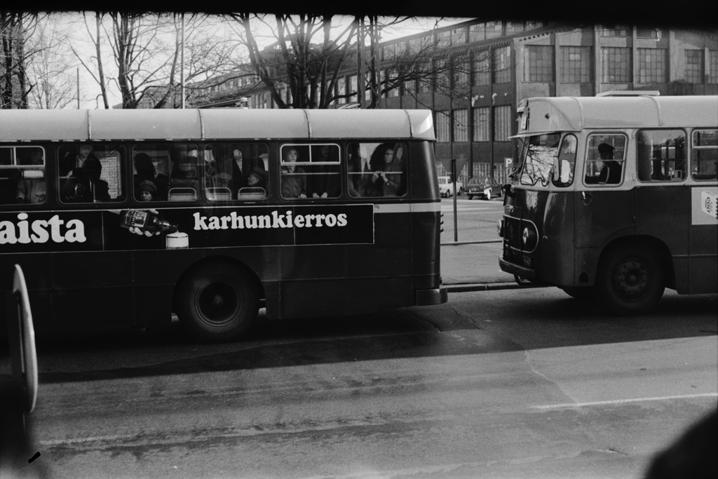 Sörnäinen, Hämeentie. Kaksi Helsingin liikennelaitoksen sinistä bussia Hämeentiellä Haapaniemenkadun kulmalla. Vasemmalla olevan täpötäyden bussin kyljessä Porin Oluttehtaan Karhu -olutmainos, oikealla olevan kyljessä Colt-savukemainos. Taustalla Oy Wärtsilä Ab:n, entisen Kone- ja Siltarakennus Oy:n teollisuusrakennus Haapaniemenkadun laidalla. Kuvattu raitiovaunun ikkunasta.