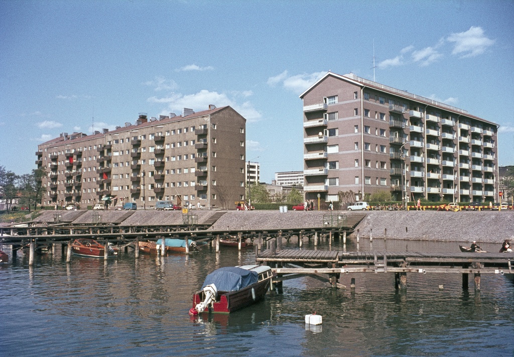 Taivallahden rantaa, Merikannontien ja Pohjoisen Hesperiankadun kulma. Etualalla laitureita ja veneitä, taustalla kerrostalot Merikannontie 3, 1 (Pohjoinen Hesperiankatu 37).