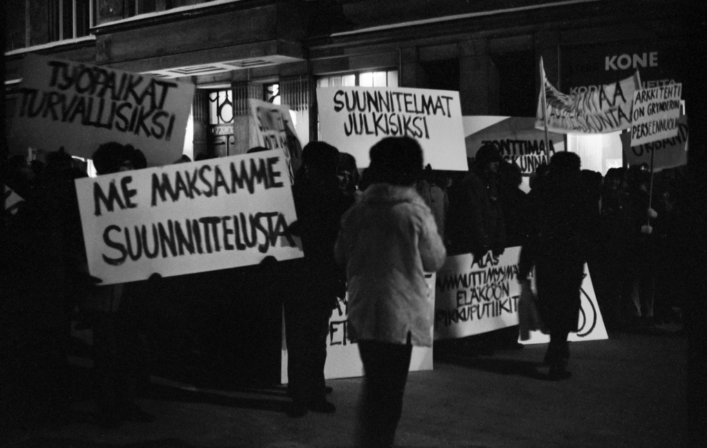 Aurorankatu 13. Mielenosoittajia plakaatteineen ja banderolleineen Aurorankatu 13. edustalla Nervanderinkadun kulmassa Taidehallissa pidettävien Suomi rakentaa 4 -näyttelyn avajaisien aikaan. Plakaateissa mm. tekstit: Me maksamme suunnittelusta ja Suunnitelmat julkisiksi.