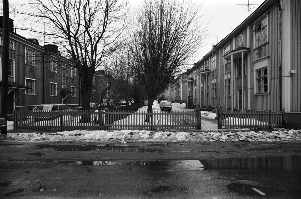 Suvannontie. Näkymä Puu-Vallilasta kevään loskaiselta Suvannontieltä kaakkoon. Vasemmalla Vanajantien 8-10 puutalojen pihapiiriä, oikealla Keuruuntien 11-13 pihapiiriä. Pihoilla parkeerattuja autoja.