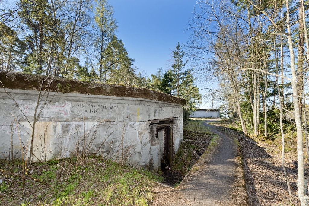 Skatanniemi. Ensimmäisen maailmansodan aikainen rannikkotykkipatteri Skatanniemessä, Vuosaaressa. Kuvassa on rintasuoja, jonka taakse on sijoitettu yhteensä neljä tykkiasemaa. Betonista valmistetun rintasuojan seinässä on ovi, josta pääsee sisälle rintasuojan alle rakennettuihin miehistö- ja varastotiloihin. Oven yläosan vahingot ovat todennäköisesti peräisin katon teräksisten tukipalkkien räjäytyksestä. Metalliset ovenkarmit on kuitenkin jätetty paikoilleen. Tiloihin johtaa pienet portaat. Oven edestä kulkee betonipäällysteinen tie, joka johtaa taustalla näkyvään seuraavaan tykkiasemaan. Kuvaussuuntana lounas.