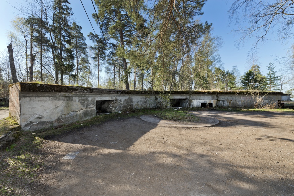 Skatanniemi. Ensimmäisen maailmansodan aikainen rannikkotykkipatteri Skatanniemessä, Vuosaaressa. Kuvassa on rintasuoja, jonka taakse on sijoitettu yhteensä neljä tykkiasemaa. Betonista valmistetun rintasuojan seinässä on kaksi ampumatarvikekomeroa. Seinällä näkyy myös kaksi pientä luukkua, joista näkee sisälle rintasuojan alle rakennettuihin miehistö- ja varastotiloihin. Rintasuojan taustalla on betonista valettu pyöreä jalusta itse tykille, jota ei koskaan ehditty asentaa. Jalustan keskellä oleva pulttirinki kertoo mihin tykki oli tarkoitus kiinnittää. Taustalla oikealla näkyy toisen tykkiaseman kaarre. Kuvaussuuntana etelä/etelä-lounas.