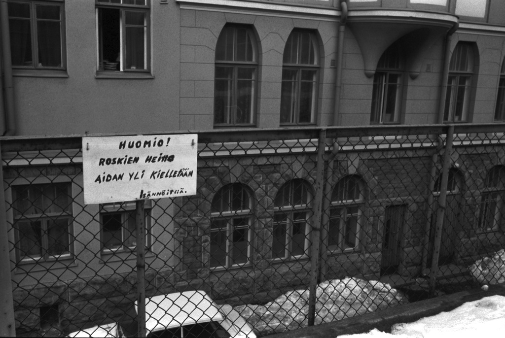 Katajanokka, Kauppiaankatu 5. Kauppiaankatu 5:n yläpihalla, varaston kattotasanteella aidassa kyltti, jossa teksti "HUOMIO! ROSKIEN HEITTO AIDAN YLI KIELLETÄÄN. ISÄNNÖITSIÄ." (sic!) Taustalla Katajanokankatu 4:n sisäpihaa.