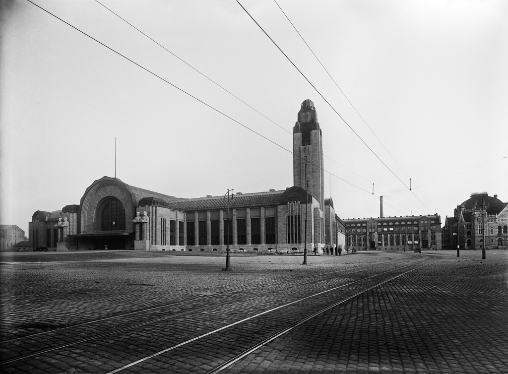Helsingin rautatieasema Kaivokadun ja Rautatientorin kulmasta nähtynä. Etualalla raitiovaunukiskot, jotka kulkevat Keskuskadun suunnalta Rautatientorille. Kellotornissa ei ole kellotaulua tai se on peitetty. Lyhdynkantaja -patsailla on pallonmuotoiset lamput käsissään, toisin kuin Sundströmin samalta paikalta talvella ottamassa kuvassa N26773. Helsingin uusi rautatieasema valmistui vuonna 1918 ja vihittiin käyttöön 1919.