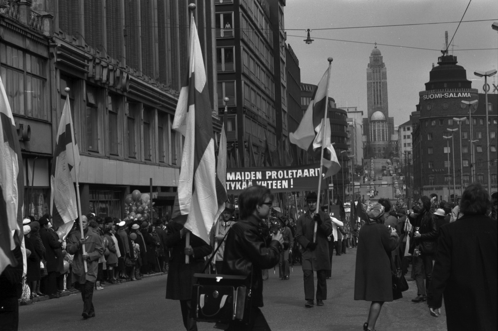 Siltasaarenkatu. Työväen vappumarssiin osallistuvia ihmisiä banderolleineen ja lippuineen kulkueessa Siltasaarenkadulla 1.5.1972. Etualalla valokuvaaja ja Suomen lippujen kantajat. Keskellä banderollissa teksti: KAIKKIEN MAIDEN PROLETAARIT, LIITTYKÄÄ YHTEEN! Taustalla Kallion kirkko.