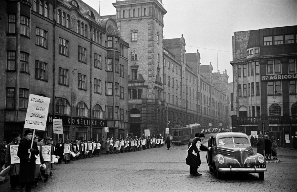 Jehovan todistaja, insinööri Kaarlo Hartevan esitelmää mainostava kulkue. Miehiä ja naisia mainos- ja tekstikylttien kanssa Rautatientorilla. Taustalla vasemmalla Suomen Koneliike Oy, oikealla matkustajakoti Agricola. Oikealla etualalla Studebaker President (vm. 1939) -henkilöauto.