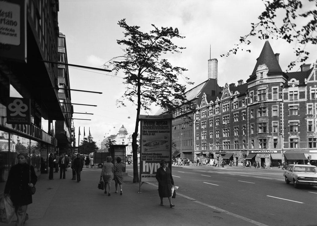 Mainospylväs Mannerheimintie 8:n kohdalla. Vasemmalla Mannerheimintie 1, Stockmannin tavaratalo. Etualalla jalankulkijoita.
