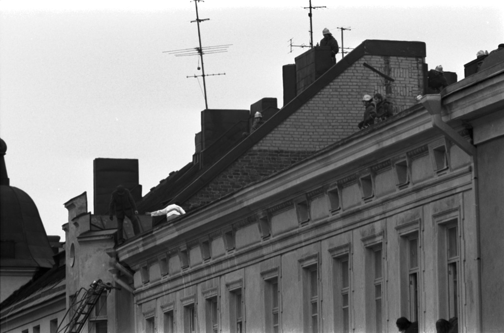 Meritullinkatu 22, 20. Meritullinkatu 22:n katolla oleva pelastusmies yrittää saada otetta Meritullinkatu 20:n katolta Meritullinkatu 22:n katolle siirtyneestä miehestä. Pelastusmies lähestyy aivan räystään reunalla seisovaa miestä. Meritullinkatu 22:n ikkunoista kurkistelee ihmisiä seuraten tapahtumaa.