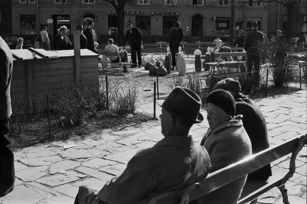 Karhupuisto. Ihmisiä istumassa penkillä Karhupuiston laidalla puistotapahtuman aikaan. Taustalla miehiä pelaamassa shakkia suuren pelilaudan äärellä.