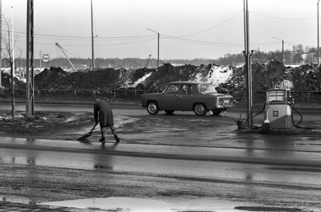 Pohjoisranta. Nainen lapioimassa loskaa Shellin huoltoasemalla Pohjoisrannassa. Taustalla likaisia lumikasoja, niiden takana Esson kyltti Tervasaarenkannaksen laidalla olevassa venesatamassa. Kuvattu Liisanpuistikon suunnalta.