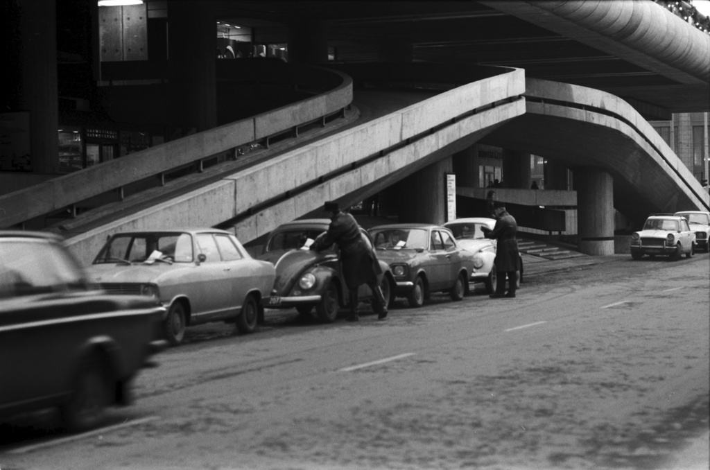Keskuskatu 6. Kaksi poliisia kirjoittamassa ja laittamassa sakkolappuja pysäköityjen autojen etuikkunaan Keskuskadulla Makkaratalon autoramppien luona.