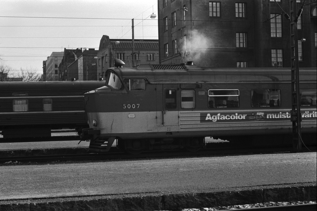 Helsingin rautatieasema. Junia Helsingin rautatieasemalla. Etualalla veturin kyljessä mainos: Agfacolor muistaa värit..