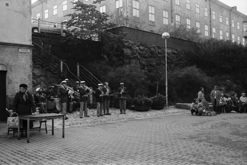 Oikokatu. Asukastapahtuma Oikokadun pihakadulla. Vasemmalla puhallinorkesteri soittamassa. Oikealla ihmisiä kerääntyneenä kirpputoripöytien ääreen. Kuvattu Kruununhaan ruotsinkielisen ala-asteen ylätasanteelta Siltavuorenpenkereen suuntaan.