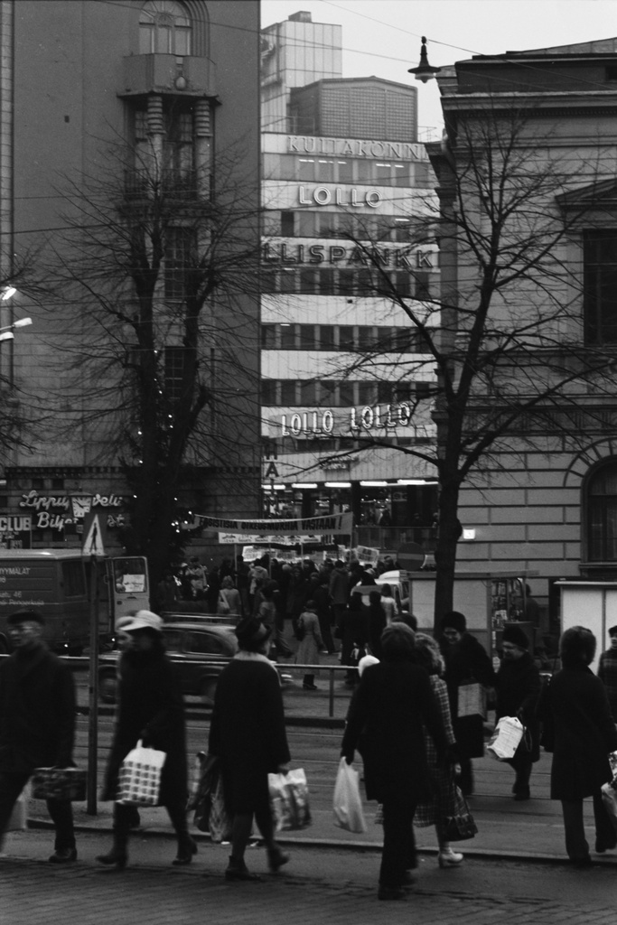 Kluuvi, Mannerheimintie. Ihmisiä ostoksineen ylittämässä katua Mannerheimintiellä. Taustalla Kaivotalo, Kaivokatu 10, ja "Ihmisoikeudet baskeille, alas Espanjan fasismi" -mielenosoituskulkue valmistautumassa liikkeellelähtöön Uuden ja Vanhan ylioppilastalon välisellä aukiolla, ns. Perunatorilla.