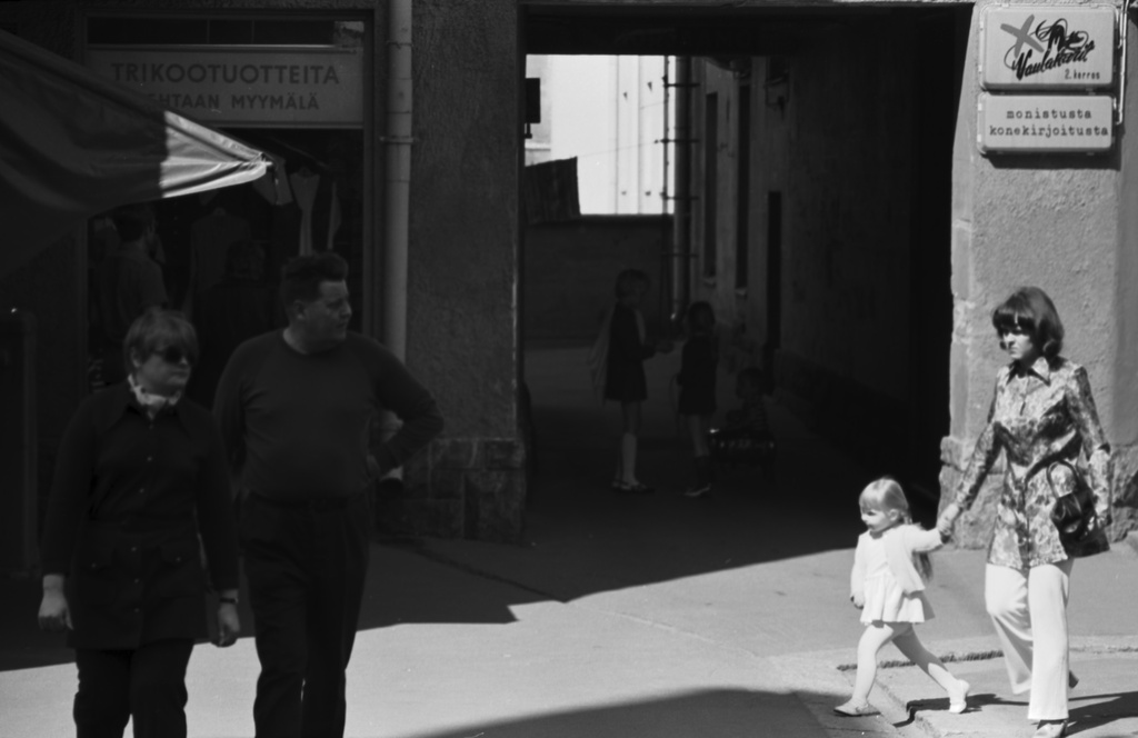 Fleminginkatu 10. Aikuisia ja lapsia Fleminginkatu 10:n edustalla. Vasemmalla trikootuotteiden tehtaan myymälä, oikealla porttikäytävän pielessä seinässä kyltti: Vaulakarit, monistusta konekirjoitusta. Porttikäytävässä kolme lasta, yksi istumassa polkuautossa.