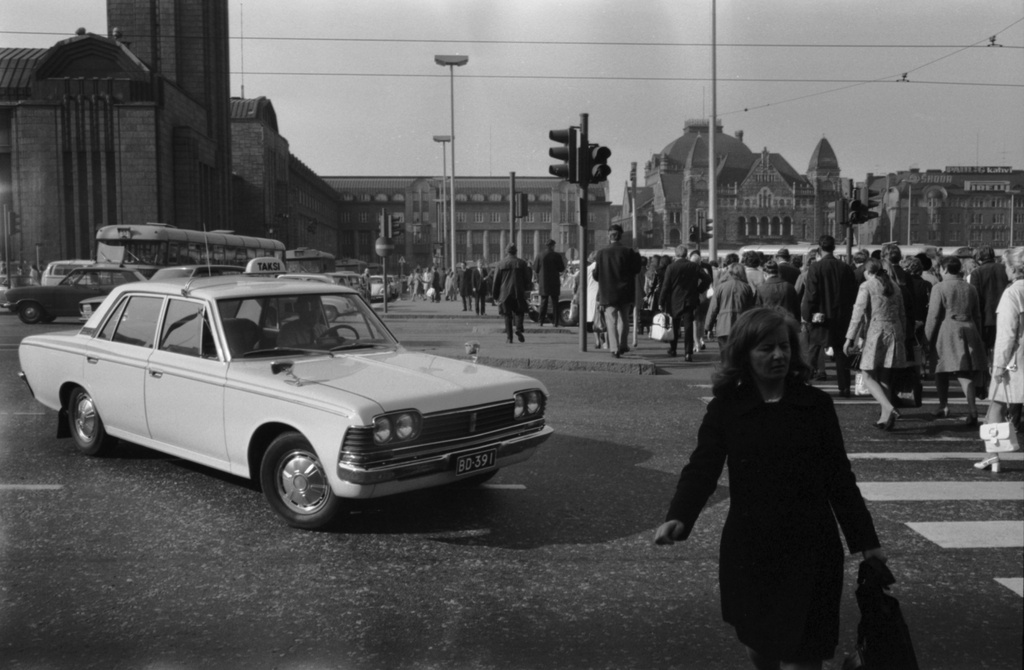 Kaivokatu. Näkymä iltapäivän ruuhka-aikaan Kaivokadulta Keskuskadun kulmasta Rautatientorin suuntaan. Vasemmalla ajoneuvoliikennettä, oikealla ihmisiä suojatiellä ylittämässä katua. Taustalla Helsingin rautatiasema sekä Kansallisteatteri. Etualan taksiauto: Toyota Crown.