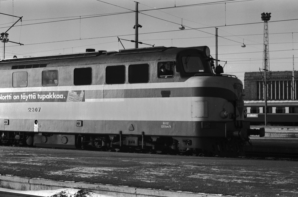 Helsingin rautatieasema. Hr12-sarjan raskas dieselveturi 2207 Helsingin rautatieasemalla. Kyljessä North State -savukemainos - Nortti on täyttä tupakkaa. Taustalla Eduskuntatalo.