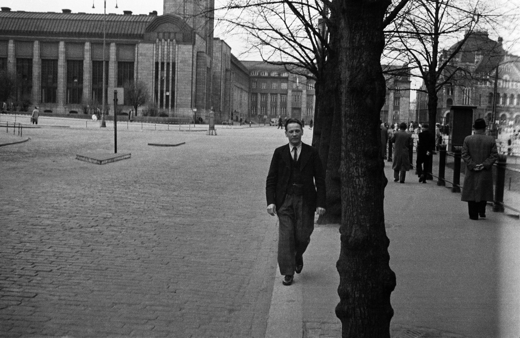 Väinö Kannisto kävelee jalkakäytävällä Keskuskadulla, taustalla Rautatieasema ja Rautatientori.