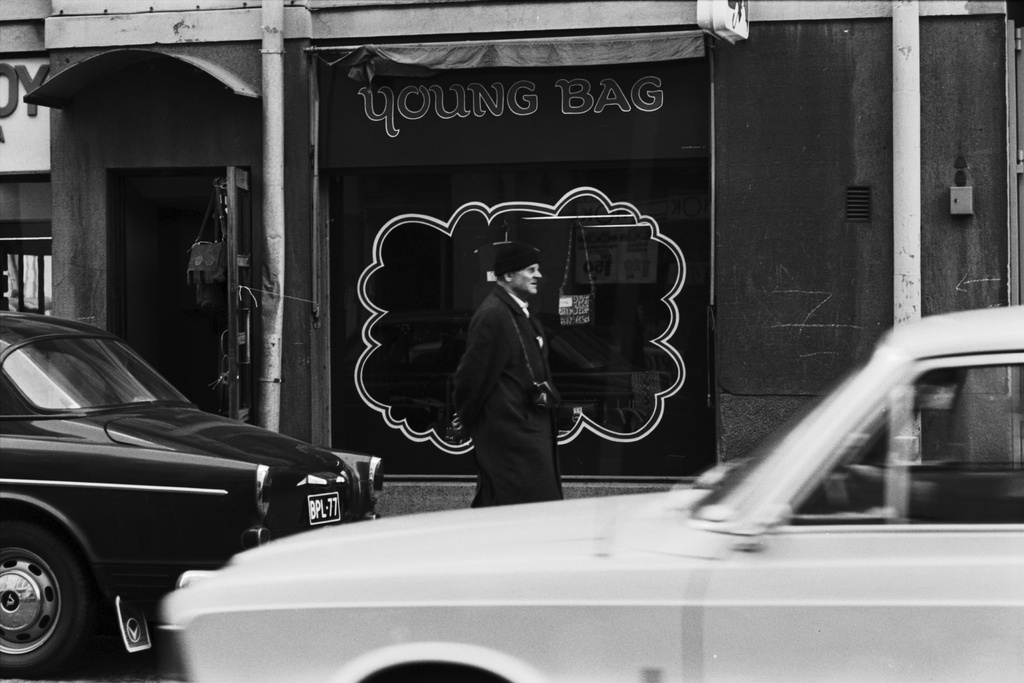 Kruununhaka, Liisankatu 15. Mies kulkemassa kamera kaulassa Liisankadulla Young bag -laukkuliikkeen näyteikkunan edessä. Kadun varressa pysäköitynä Volvo Amazon Favorit -henkilöauto.