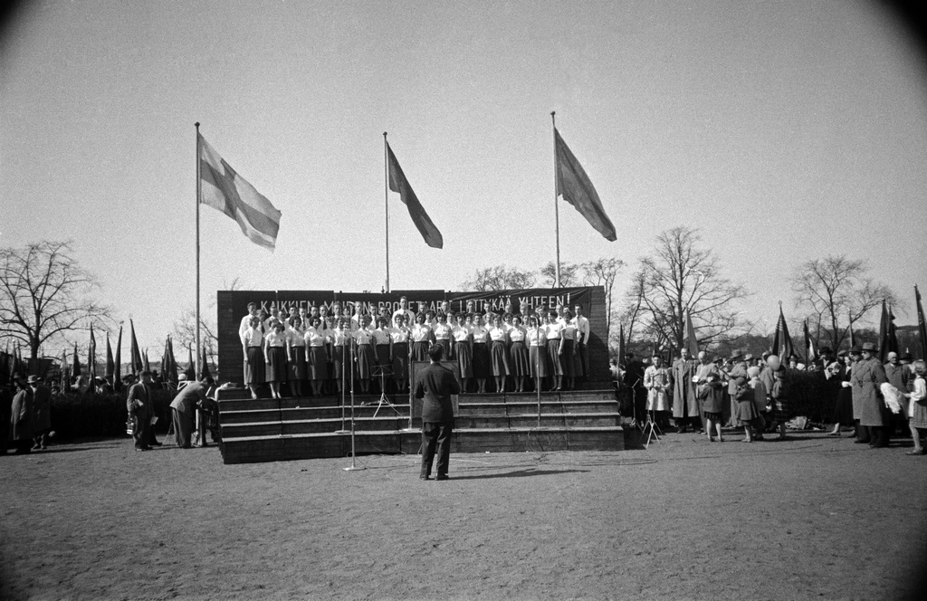 Työväen vappujuhla Kaisaniemessä. Koiton Nuorisokuoro esiintyy. Taustalla Suomen, Neuvostoliiton ja Ruotsin liput sekä teksti "Kaikkien maiden proletaarit, liittykää yhteen!".