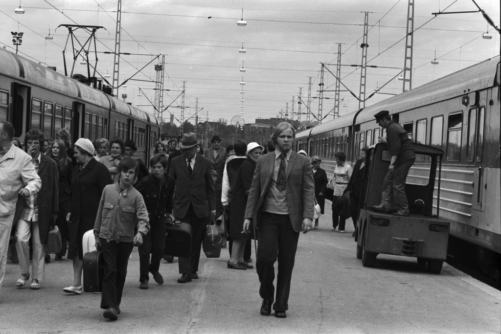 Helsingin rautatieasema. Asemalle saapuneista junista tulevia ihmisiä matkalaukkuineen asemalaiturilla. Vasemmalla lähiliikenteen sähköjuna. Oikealla kaukoliikenteen dieselmoottorijuna, jonka vieressä laituriauto.
