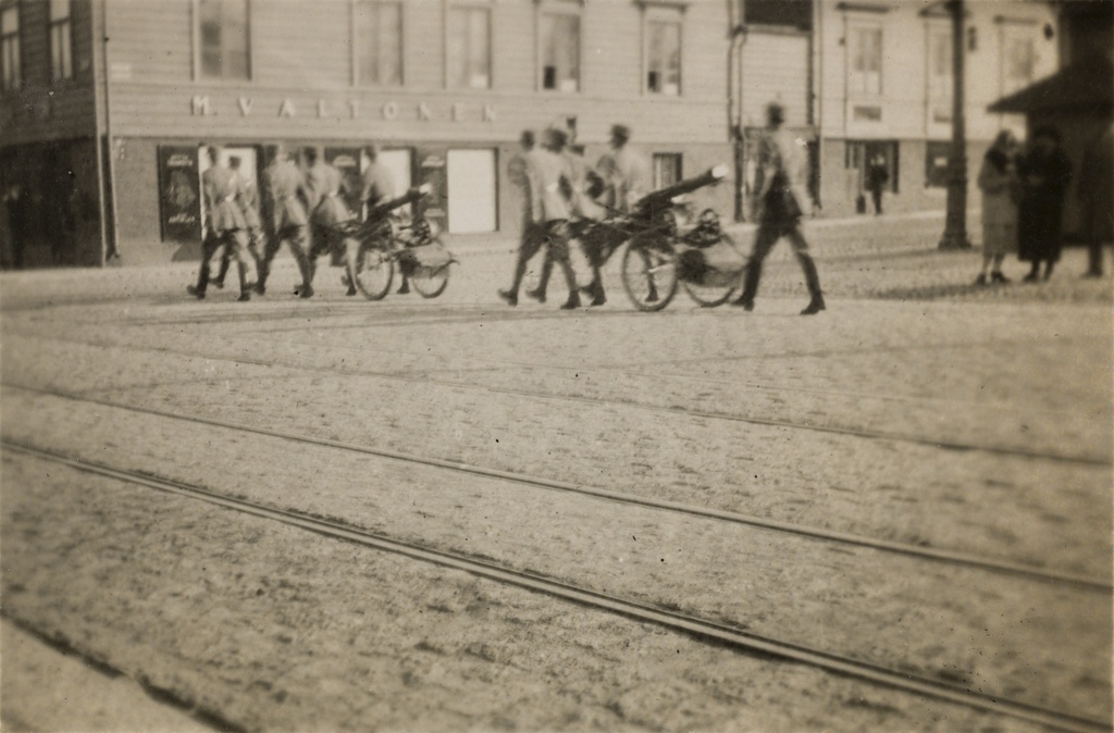 Sotilaita kuljettamassa konekiväärejä, taustalla Läntinen Heikinkatu 26, myöhemmin Mannerheimintie 20, jossa toimi M. Valtosen herrain vaatetus- ja kangaskauppa.