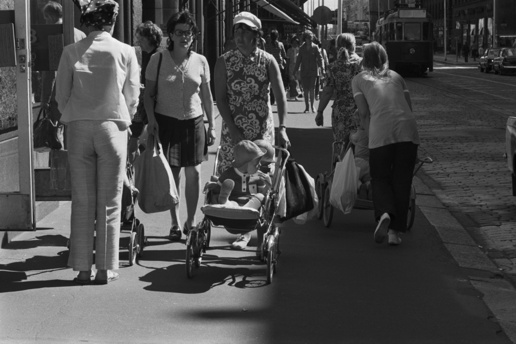 Aleksanterinkatu. Jalankulkijoita aurinkoisella Aleksanterinkadulla. Mm. kaksi naista kukkaleningeissään sekä lastenrattaita työntäviä naisia.